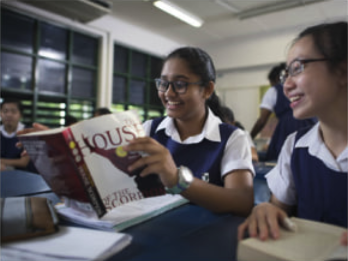 Two students in uniform smiling and looking at a book together within a classroom setting. 